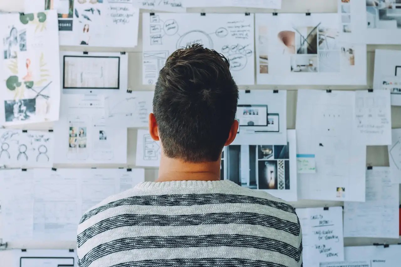 A man, seen from behind, looks at a wall covered with pinned documents, diagrams and notes