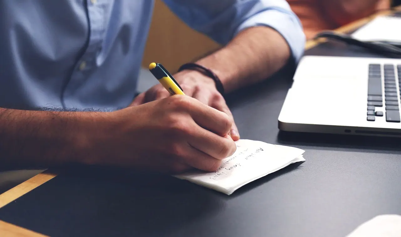 A hand takes notes with a pen on a sheet of paper next to a laptop