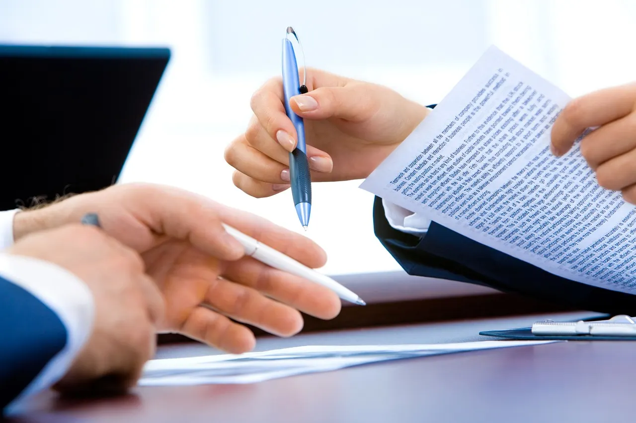 Two people proofreading a printed document, pens in hand, next to a laptop
